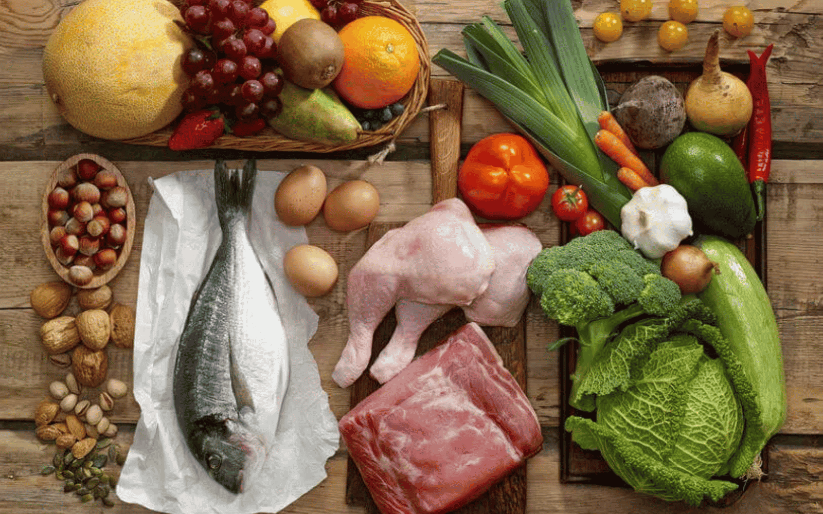 Różnorodne produkty spożywcze ułożone na drewnianym stole, w tym ryba, mięso, jajka, warzywa, owoce, orzechy i nasiona, przedstawiające zbilansowaną dietę wspierającą prawidłowe funkcjonowanie wątroby oraz dostarczającą niezbędnych składników odżywczych.