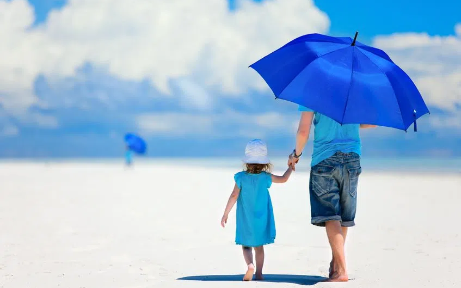 Dwie osoby idące po jasnej, piaszczystej powierzchni pod otwartym niebem, jedna osoba trzyma nad sobą niebieski parasol, druga osoba w jasnym stroju trzyma ją za rękę