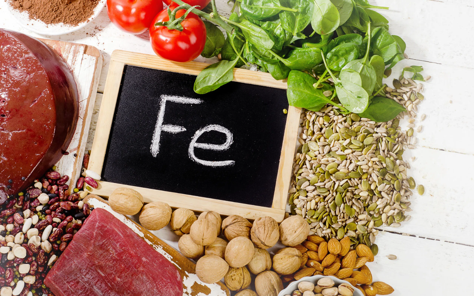 Aranżacja produkt&oacute;w spożywczych ułożonych na jasnej powierzchni: zielone liście szpinaku, pestki dyni, orzechy, migdały, rośliny strączkowe, czerwone mięso, pomidory oraz tabliczka z napisem &bdquo;Fe&rdquo;