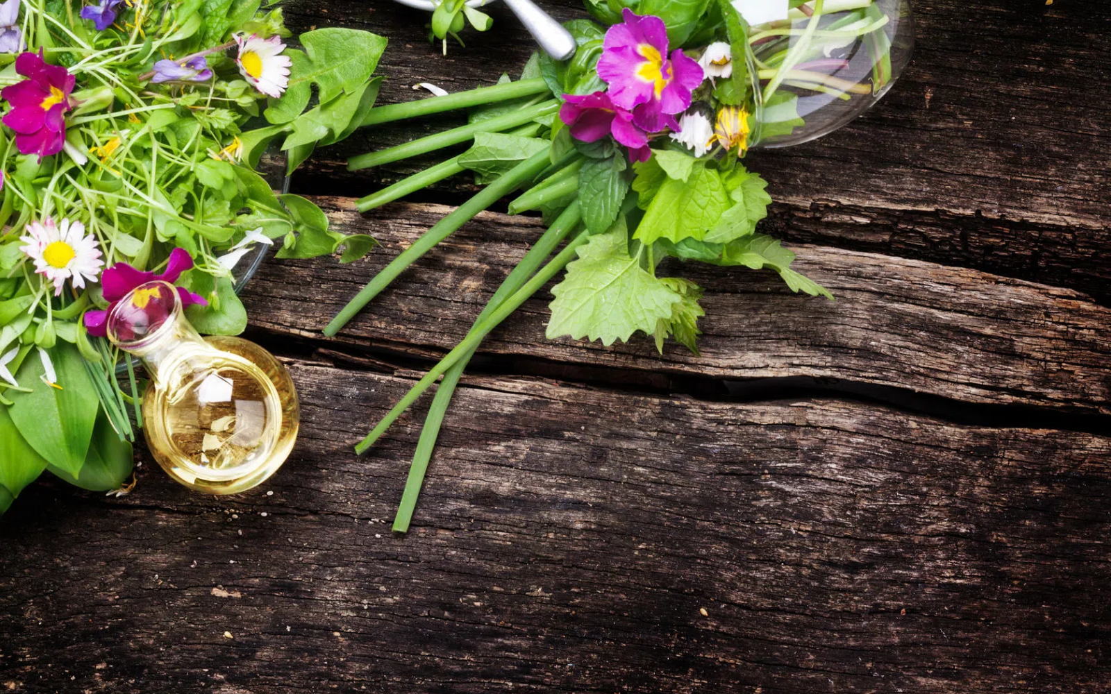 Kompozycja świeżych i suszonych zi&oacute;ł oraz jadalnych kwiat&oacute;w ułożonych na ciemnej, drewnianej powierzchni, obok mała butelka z jasnym olejem roślinnym i metalowa łyżka