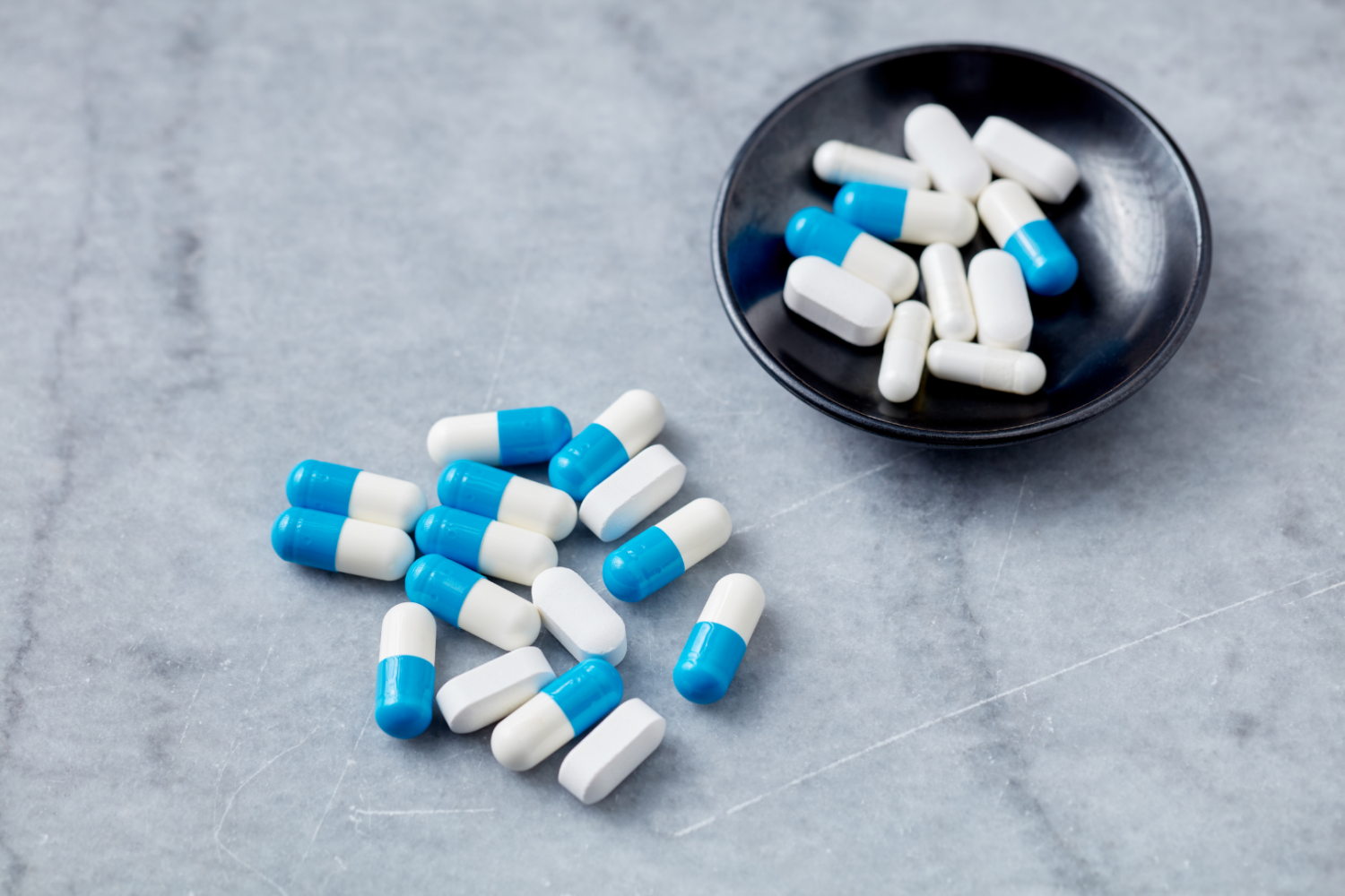 Białe i niebieskie kapsułki oraz tabletki rozsypane na szarej powierzchni obok czarnej miseczki wypełnionej kapsułkami