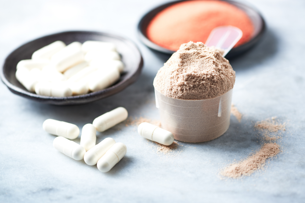 Miarka z brązowym proszkiem beta-alaniny na marmurowym blacie, obok rozsypane białe kapsułki i czarna miseczka z kapsułkami, w tle pomarańczowy proszek w miseczce