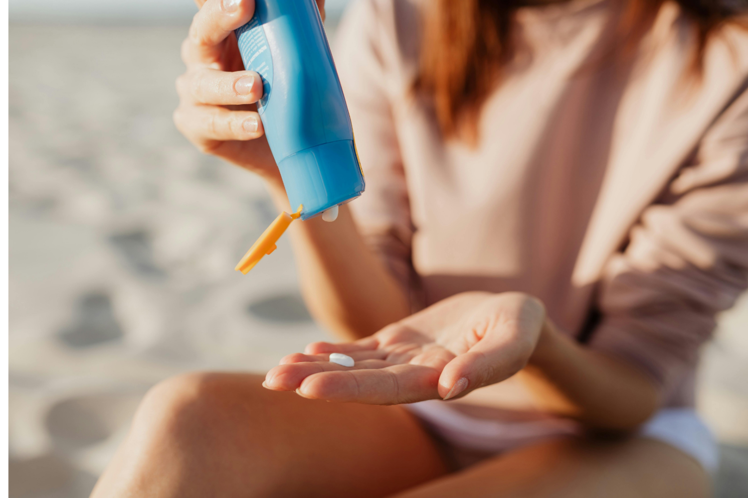 Dłoń trzymająca niebieską butelkę z kremem, niewielka ilość preparatu wyciskana na otwartą dłoń, w tle piaszczysta plaża
