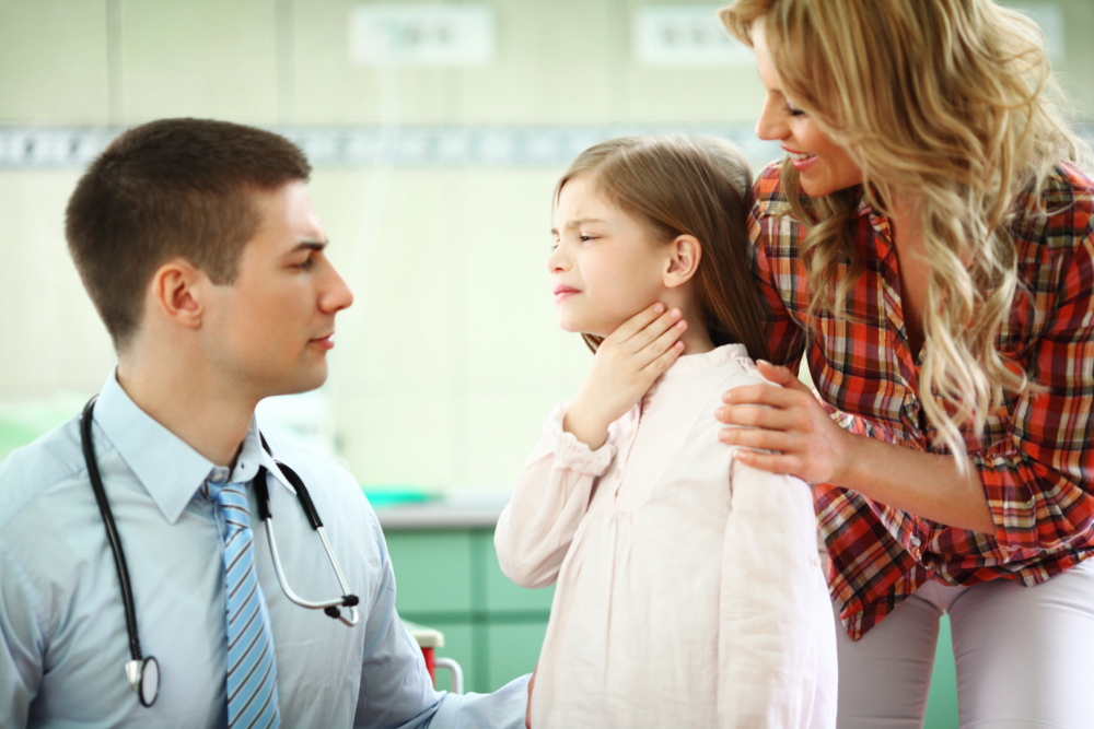 Dziewczynka trzymająca dłoń na szyi podczas wizyty u lekarza, obok osoba dorosła wspierająca dziecko