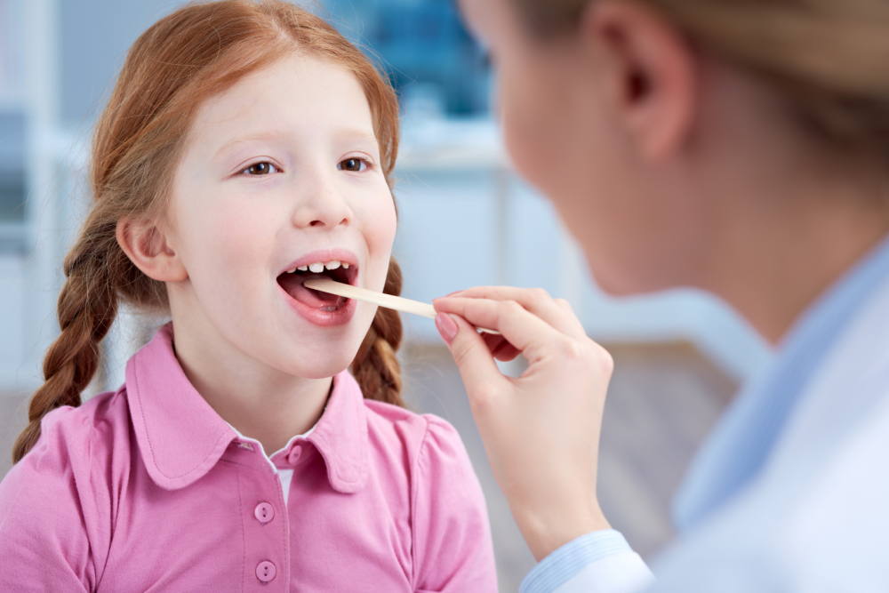 Dziecko w różowej koszulce podczas badania gardła drewnianą szpatułką przez lekarza