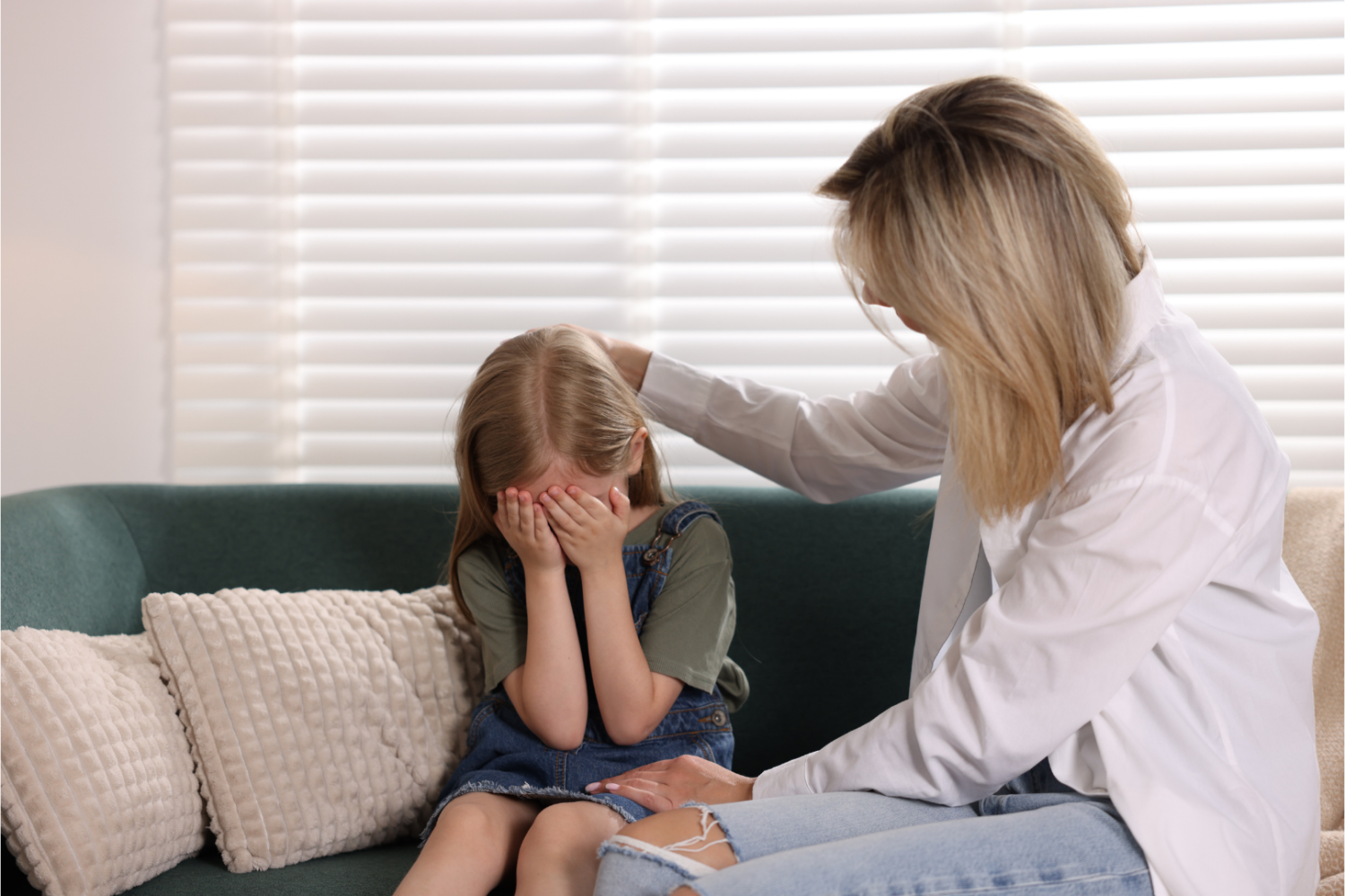 Dziecko siedzące na kanapie z pochyloną głową i dłońmi zasłaniającymi twarz, obok osoba dorosła kładąca rękę na głowie dziecka