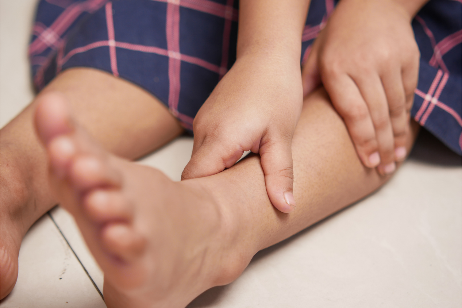 Dziecko siedzące na podłodze, trzymające dłonią okolice podudzia i kostki, ujęcie z bliska na nogi i ręce