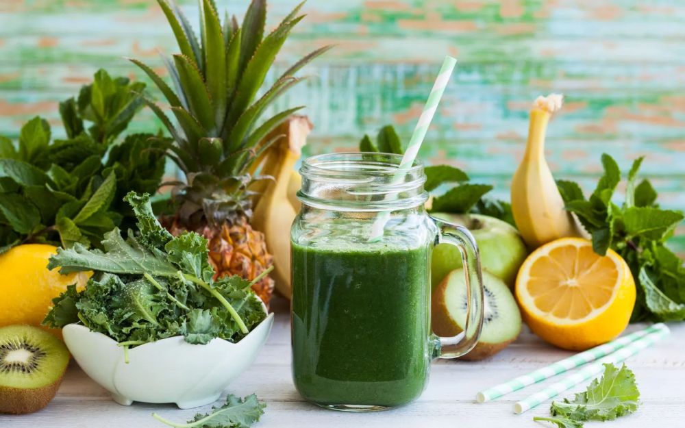 Szklany słoik z zielonym koktajlem na bazie spiruliny i słomką, otoczony świeżymi owocami i warzywami: ananasem, bananem, kiwi, cytryną, jarmużem i miętą