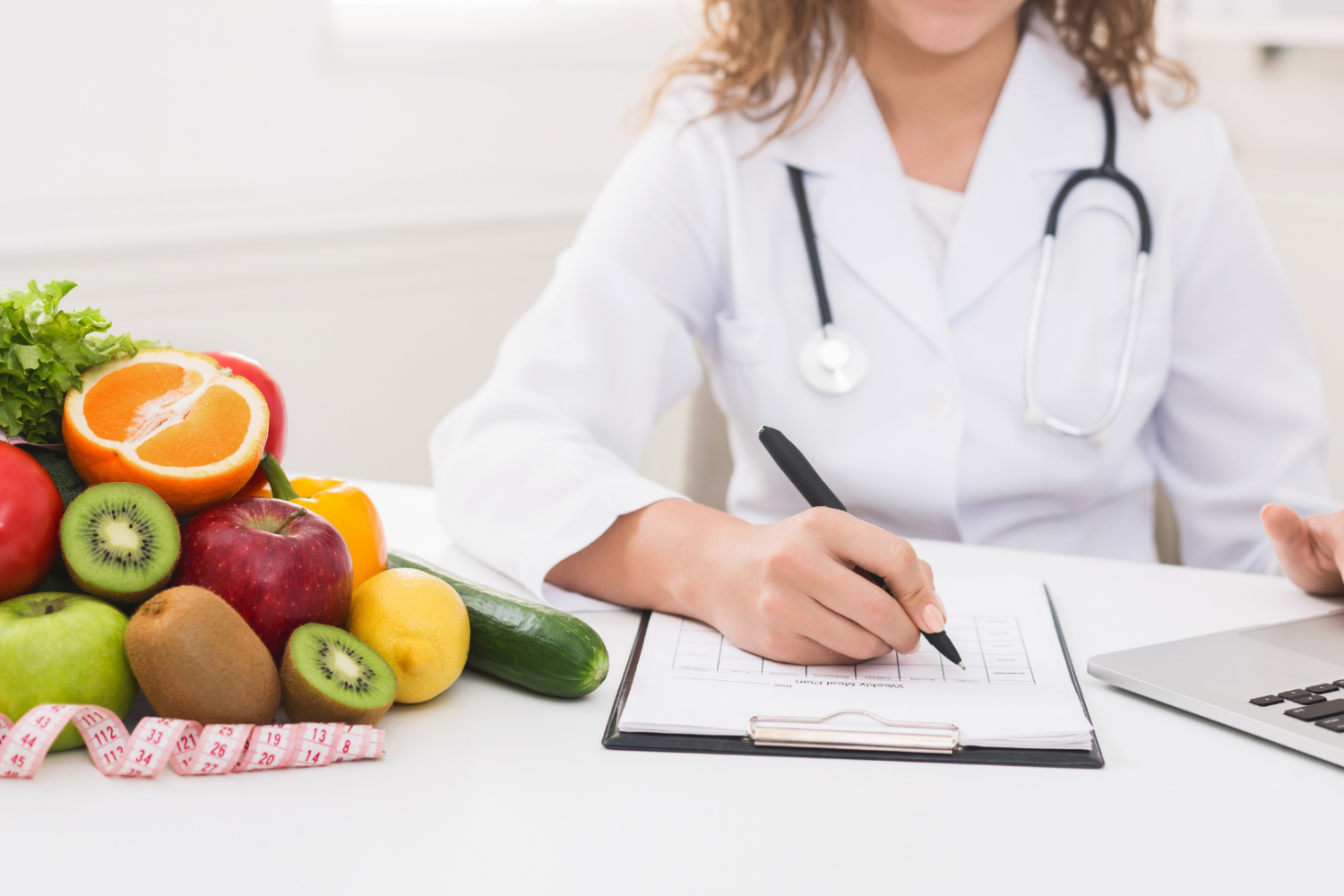 Osoba w białym fartuchu siedząca przy biurku, zapisująca notatki w formularzu, obok laptop oraz zestaw świeżych owoców, warzyw i miarka krawiecka