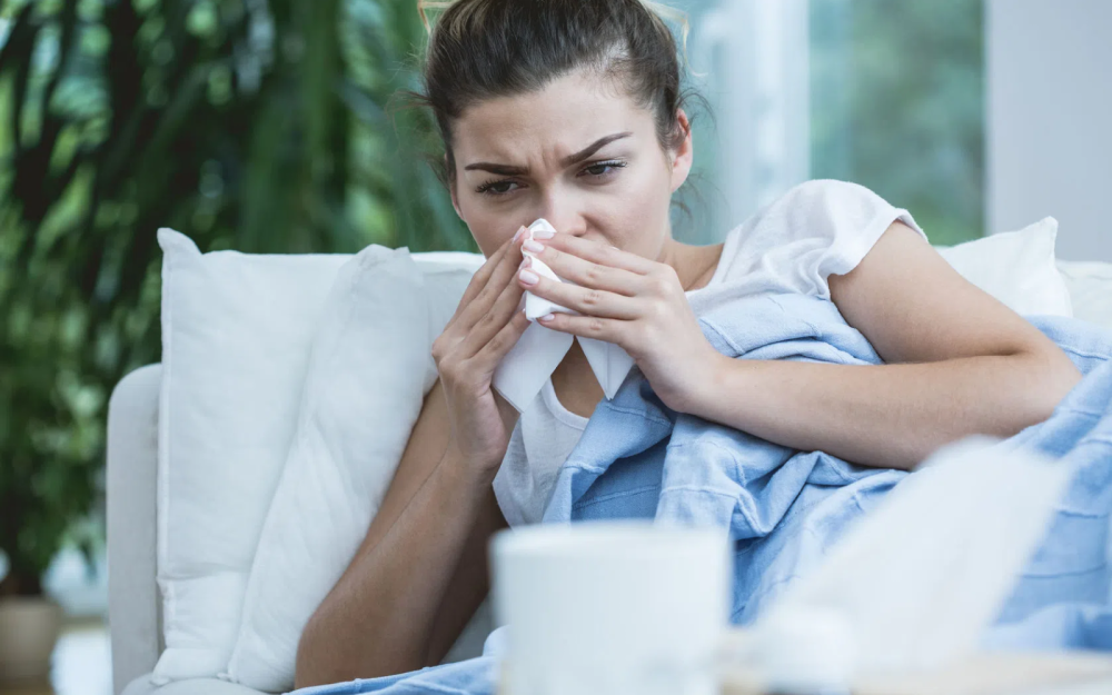 Osoba siedząca na kanapie owinięta niebieskim kocem, trzymająca chusteczkę higieniczną w dłoniach, na stoliku w tle biały kubek