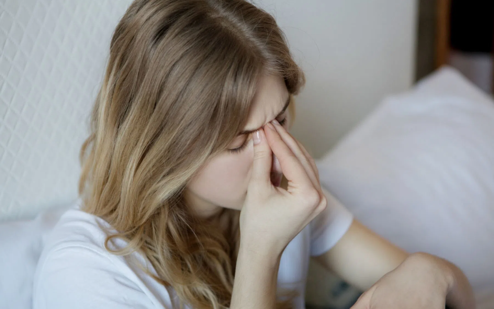 Osoba siedząca na łóżku w jasnym pomieszczeniu, ubrana w białą koszulkę, trzymająca chusteczkę przy nosie