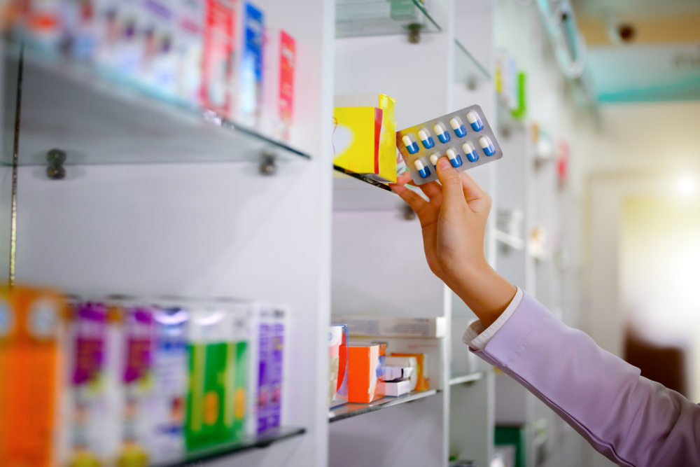Dłoń sięgająca po blister z kapsułkami z półki aptecznej, w tle regały z opakowaniami leków