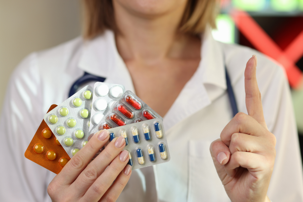 Osoba w białym fartuchu trzymająca kilka blistrów z tabletkami i kapsułkami, druga dłoń uniesiona z wyprostowanym palcem