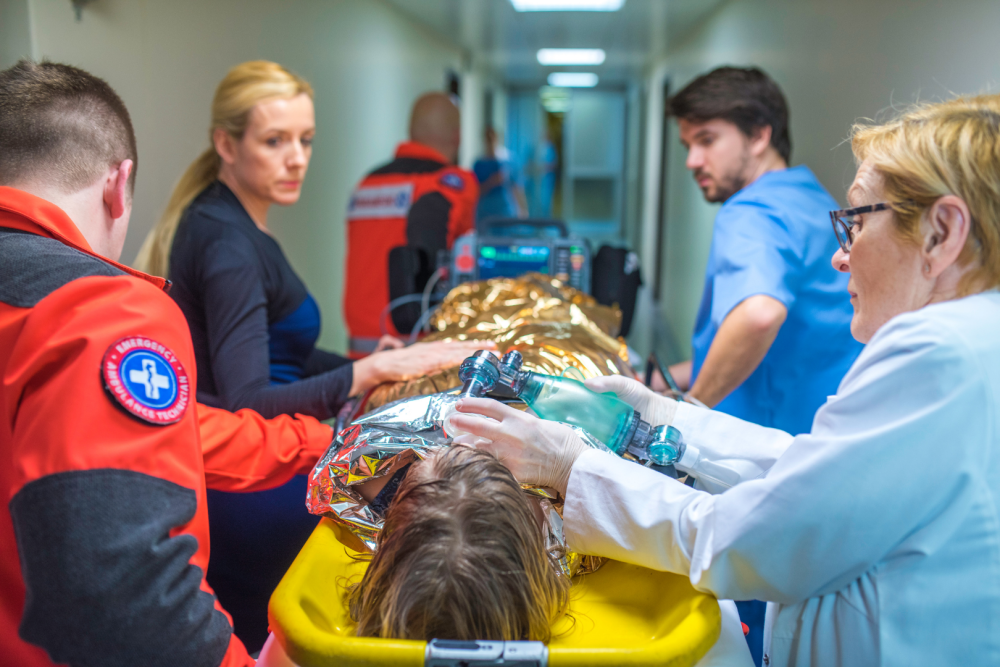 Zespół ratowników medycznych transportujący osobę okrytą folią termoizolacyjną na noszach, jeden z ratowników wykonuje wentylację workiem samorozprężalnym