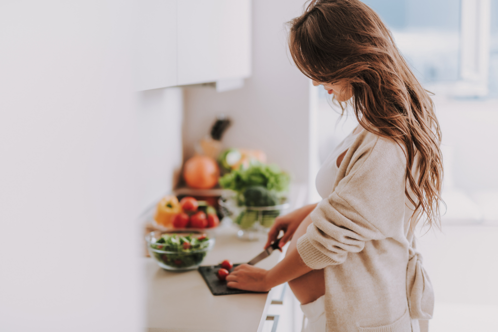 Osoba krojąca świeże warzywa na desce w jasnej kuchni, obok miski sałatki i kolorowych papryk