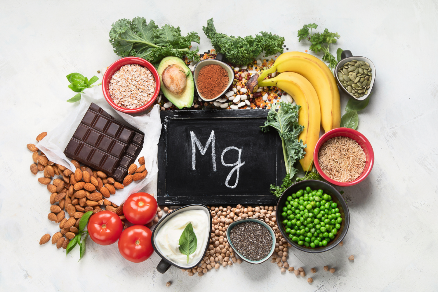 Produkty spożywcze bogate w magnez ułożone wokół tabliczki z napisem „Mg”, w tym banany, orzechy, zielone warzywa liściaste, nasiona i kakao, przedstawiające naturalne źródła magnezu w diecie.