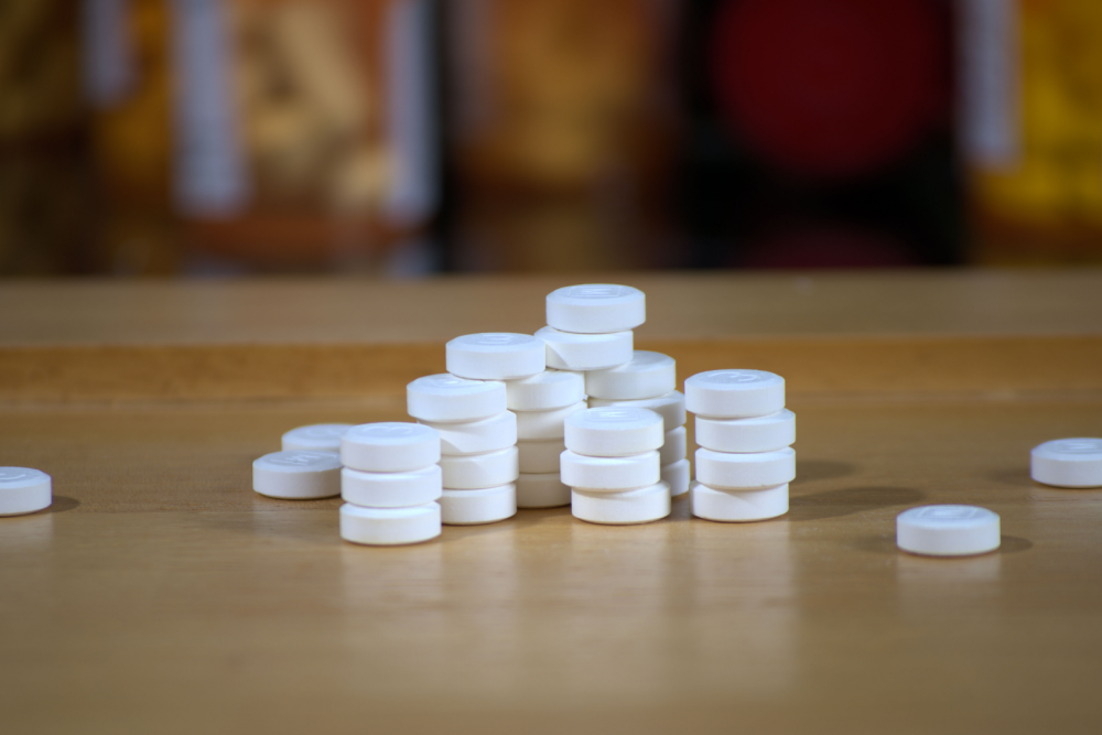 Stos białych tabletek ułożonych na drewnianym blacie, w tle rozmyte kolorowe opakowania