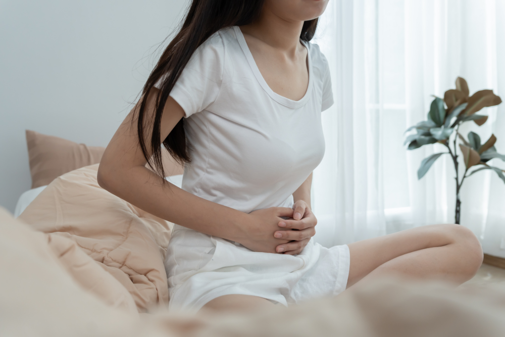 Osoba siedząca na łóżku w jasnym pokoju, ubrana w białą koszulkę, trzymająca dłonie na brzuchu