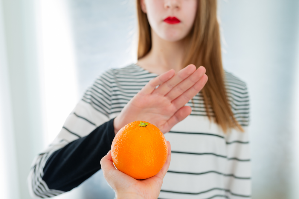 Osoba w pasiastym swetrze odmawiająca przyjęcia pomarańczy, trzymając dłoń w geście stop