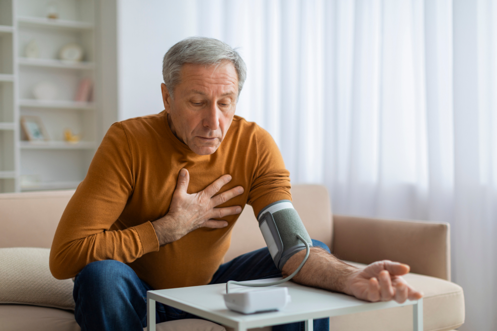 Mężczyzna w średnim wieku siedzący na kanapie w domu, trzymający rękę na klatce piersiowej, z mankietem ciśnieniomierza założonym na przedramieniu