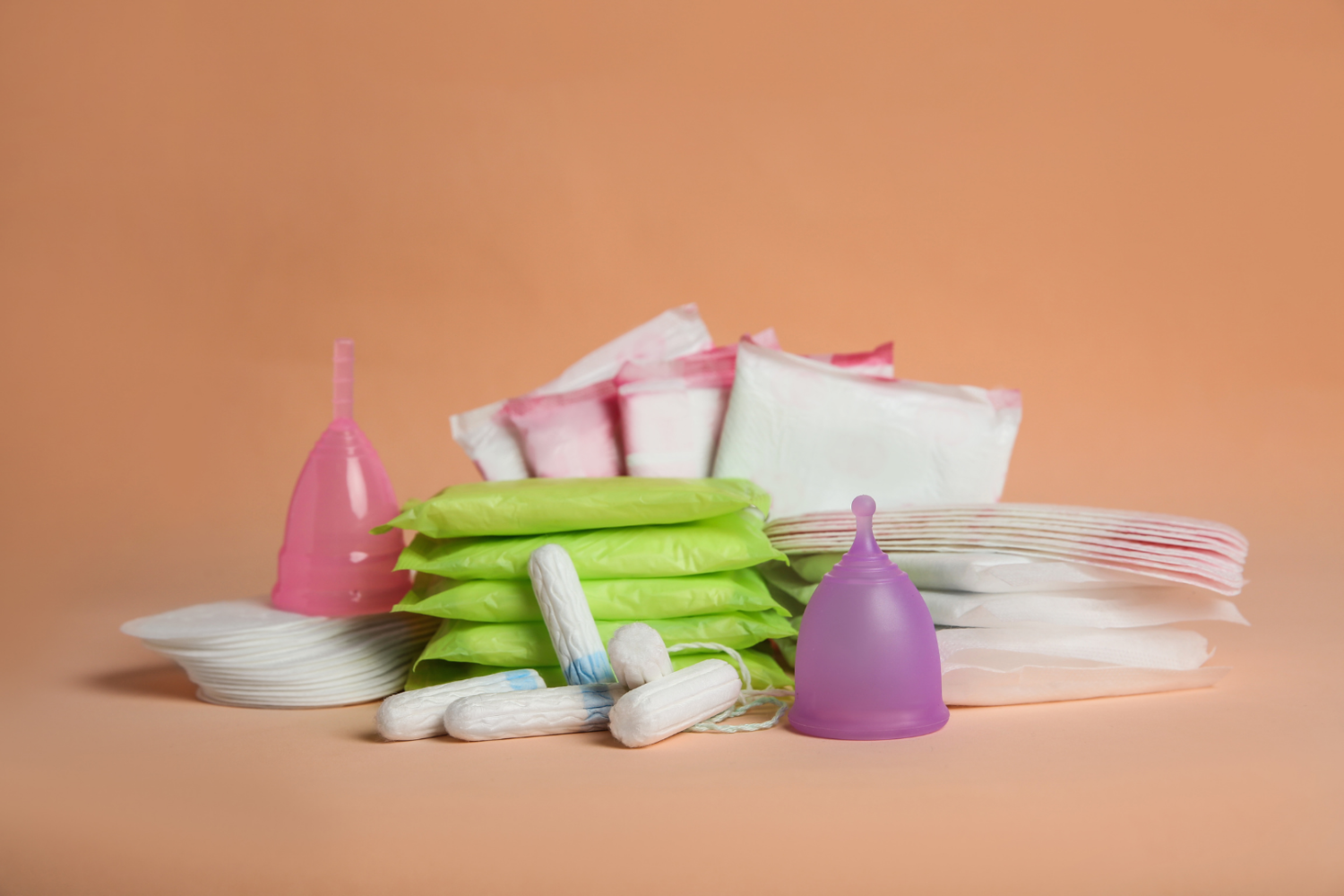 Na jednolitym tle ułożone różne środki higieny menstruacyjnej: podpaski, tampony oraz kubeczki menstruacyjne w dwóch kolorach