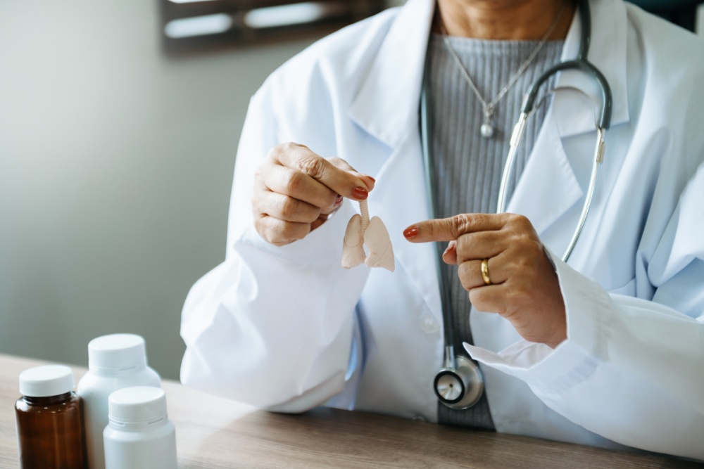 Lekarz w białym fartuchu trzymający papierowy model płuc i wskazujący na jego elementy, na stole stoją brązowe butelki z lekami