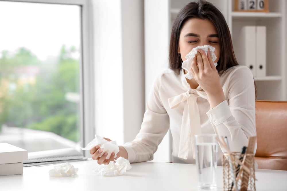 Osoba siedząca przy biurku w jasnym pomieszczeniu, trzymająca chusteczkę higieniczną w dłoni, na blacie szklanka wody i pudełko chusteczek