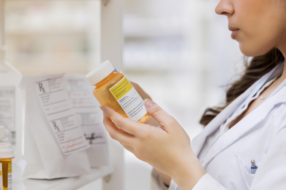 Osoba w białym fartuchu trzymająca w dłoniach pomarańczową butelkę z lekiem i czytająca etykietę, w tle półki apteczne oraz dokumenty