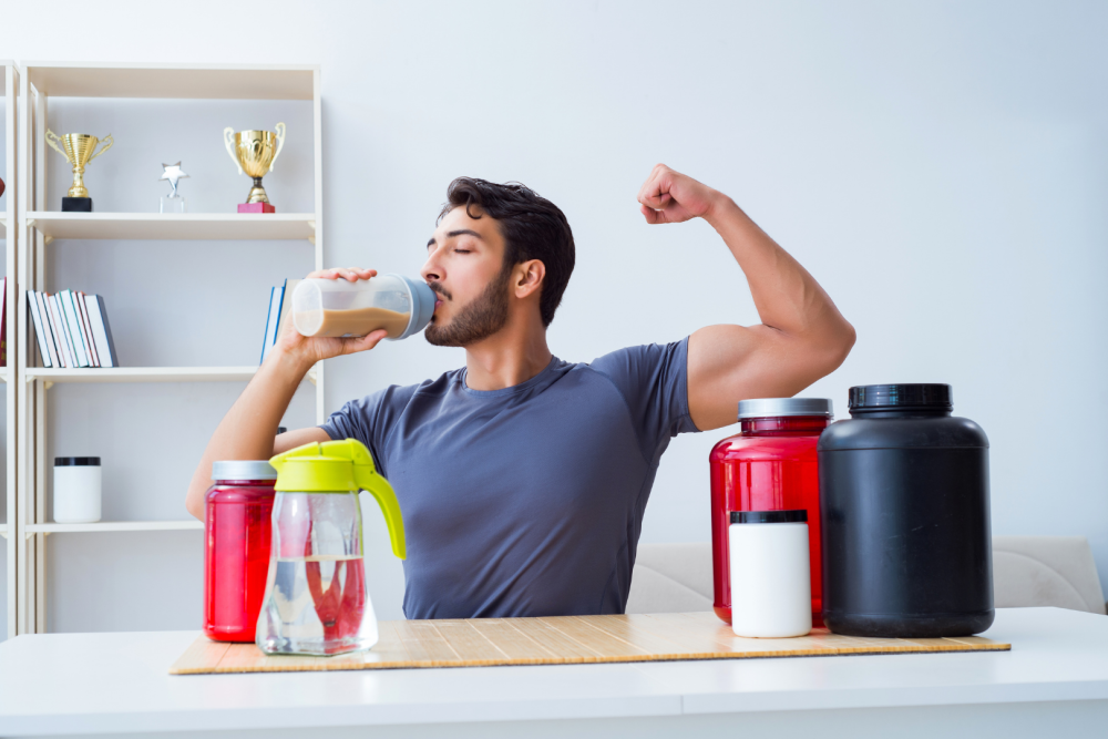Osoba pijąca napój z shakera, siedząca przy stole z pojemnikami na suplementy i akcesoriami treningowymi