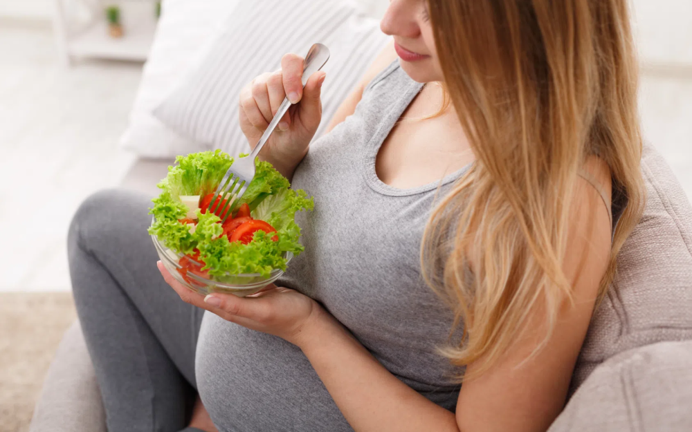 Osoba w ciąży siedząca na kanapie, trzymająca miskę z sałatką z zielonej sałaty i pomidorów oraz widelec