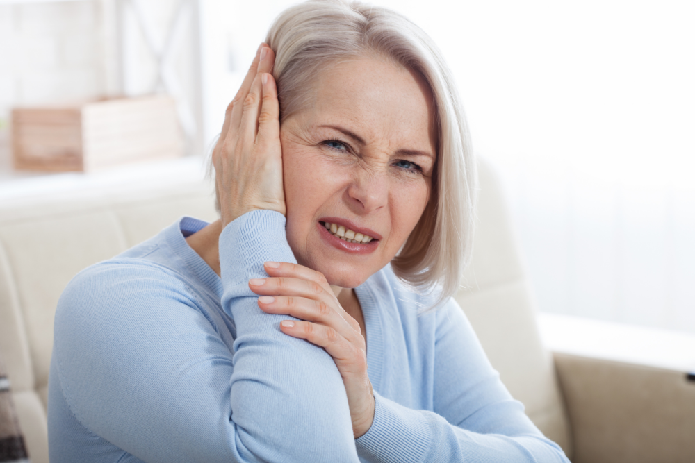 Osoba siedząca na kanapie w jasnym wnętrzu, ubrana w niebieski sweter, trzymająca dłonie przy uchu