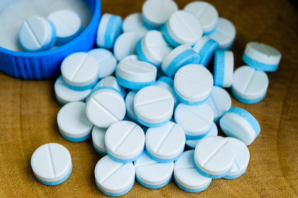 Niebiesko‑białe tabletki rozsypane na drewnianej powierzchni, obok niebieska zakrętka od opakowania
