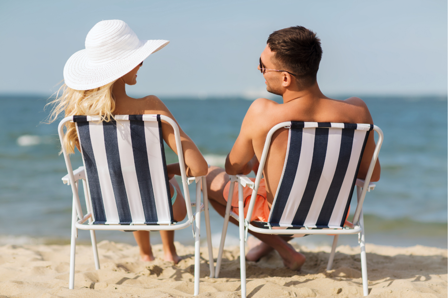 Dwie osoby siedzące na składanych leżakach na piaszczystej plaży, w tle morze i jasne niebo, jedna osoba ma szerokie nakrycie głowy