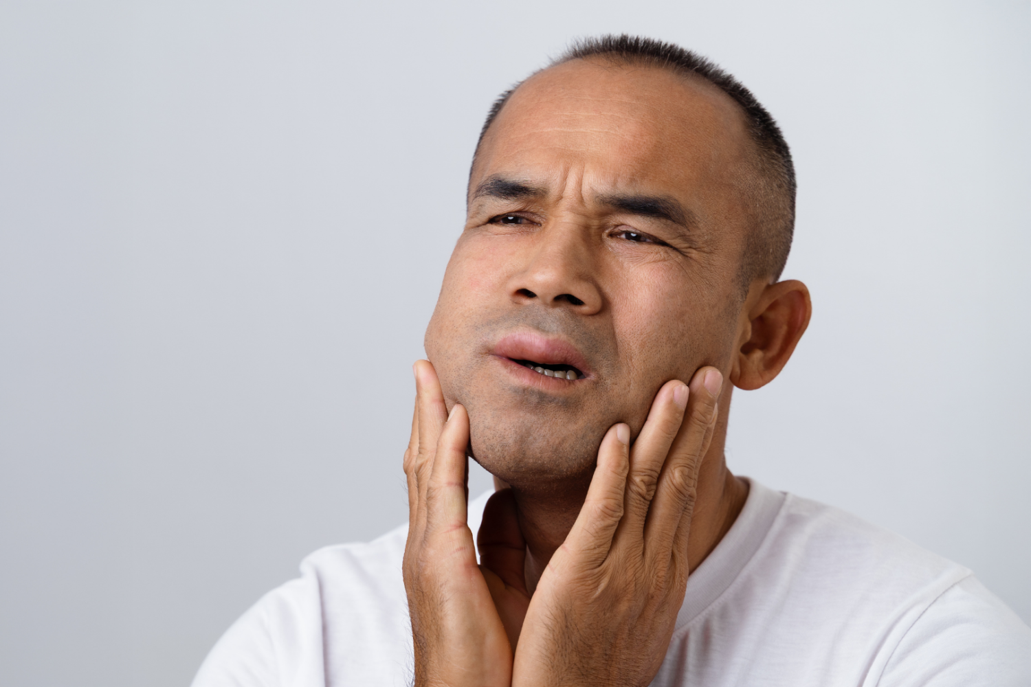 Osoba dotyka dłonią okolicy żuchwy i szyi, usta lekko otwarte, tło jednolite