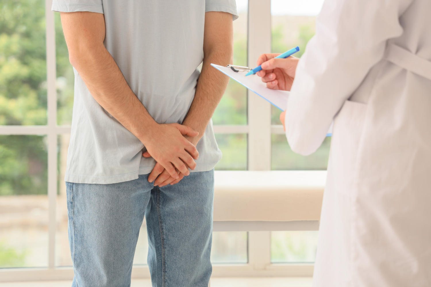 Konsultacja w gabinecie lekarskim, osoba stojąca z dłońmi złożonymi z przodu, druga osoba trzyma clipboard i długopis, w tle jasne wnętrze z oknem