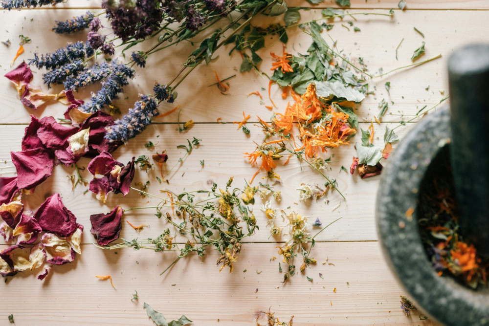 Drewniany st&oacute;ł z rozsypanymi suszonymi ziołami: płatki r&oacute;ż, kwiaty nagietka, lawenda i liście mięty, obok moździerz z tłuczkiem