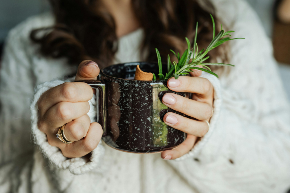 Osoba w jasnym swetrze trzymająca w dłoniach ceramiczny kubek z naparem ziołowym, ozdobiony gałązką rozmarynu i laską cynamonu