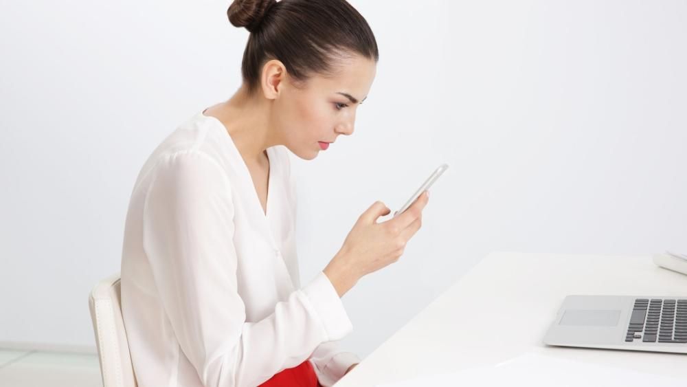 Kobieta siedząca przy biurku z lekkim garbem spowodowanym patrzeniem w dół w telefon