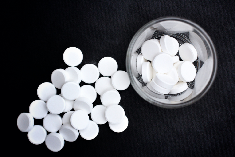 Białe, okrągłe tabletki rozsypane na czarnym tle oraz szklany pojemnik wypełniony tabletkami
