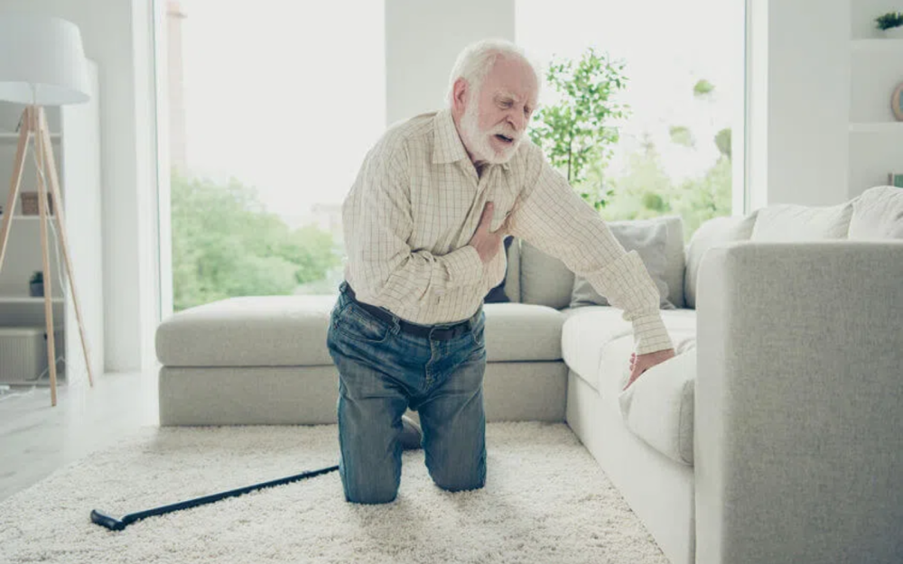 Osoba klęcząca na podłodze w jasnym salonie, jedną dłonią opierająca się o sofę, drugą trzymająca się za klatkę piersiową