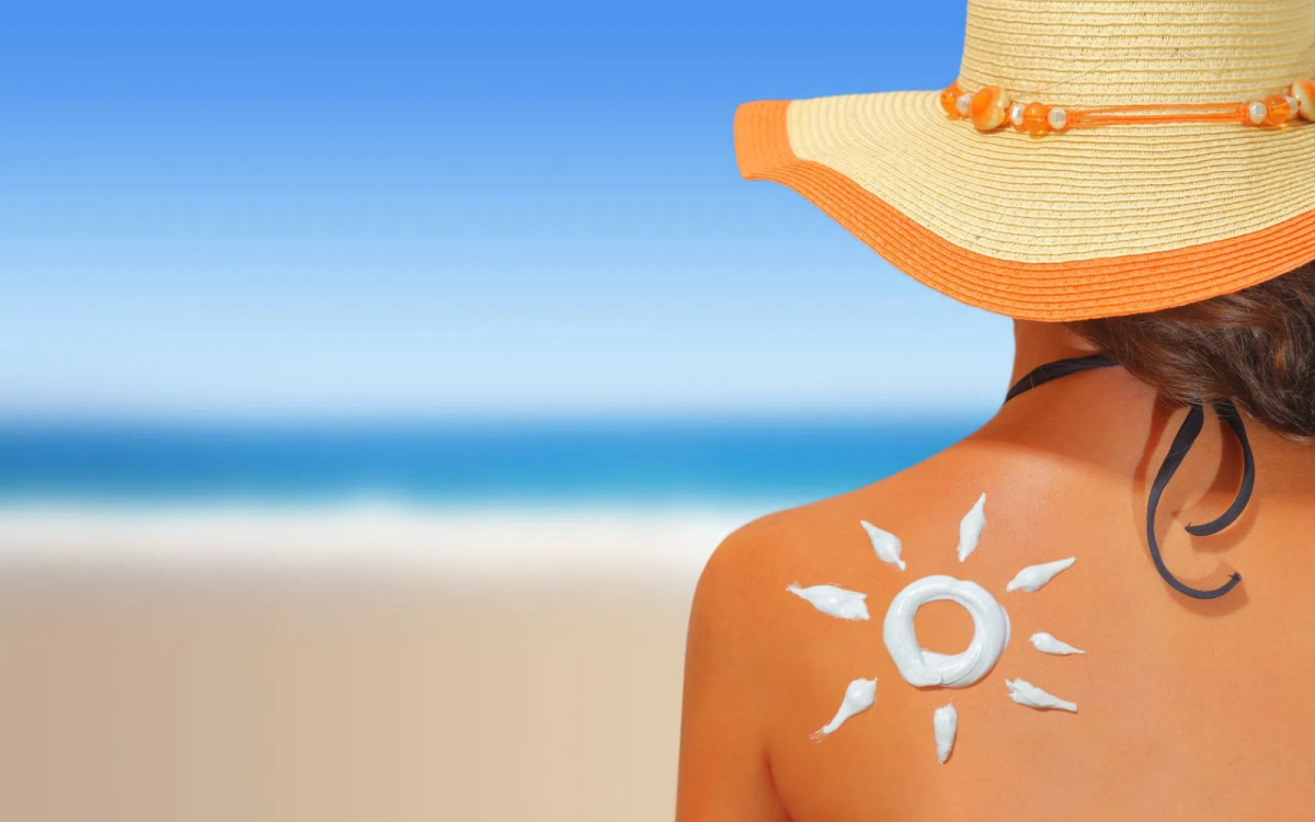 Plecy osoby na plaży z kapeluszem przeciwsłonecznym, na skórze ramienia widoczny krem ochronny z filtrem UV aplikowany w kształcie słońca, symbolizujący ochronę przed promieniowaniem słonecznym i pielęgnację skóry.