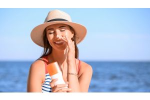 Uśmiechnięta kobieta w kapeluszu na plaży trzyma krem SPF