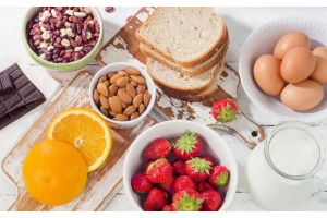 Różnorodne produkty spożywcze ułożone na stole: truskawki, pomarańcza, migdały, mieszanka fasoli, jajka, ciemna czekolada, kromki chleba oraz szklany dzbanek mleka