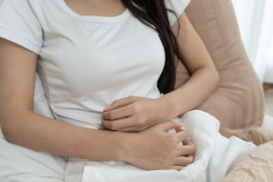 Osoba siedząca na łóżku w jasnym wnętrzu, trzymająca obie dłonie na dolnej części brzucha