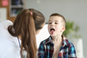 Lekarka pochylająca się nad dzieckiem w koszuli w kratę podczas badania gardła w gabinecie