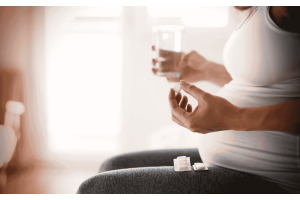 Osoba w ciąży siedząca na kanapie, ubrana w jasny top, trzymająca w jednej dłoni szklankę wody, a w drugiej tabletkę, na kolanach opakowanie leków