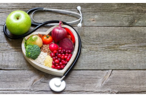 Drewniany stół z misą w kształcie serca wypełnioną zdrowymi produktami: brokułem, pomidorem, cebulą, burakiem, kaszą i żurawiną, obok zielonego jabłka i stetoskopu