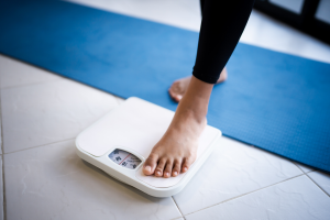 Stopa stawiana na mechanicznej wadze łazienkowej stojącej na jasnej podłodze, w tle mata do ćwiczeń