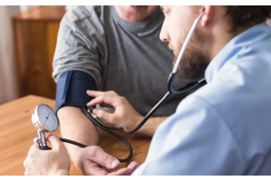 Pomiar ciśnienia krwi wykonywany w domu lub gabinecie – osoba siedząca przy stole z mankietem ciśnieniomierza na ramieniu, druga osoba kontroluje wynik za pomocą aparatu i stetoskopu.