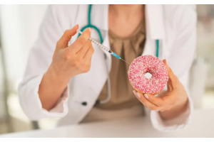 Dłonie osoby w białym fartuchu trzymające różowego pączka posypanego cukrem oraz strzykawkę skierowaną w jego stronę